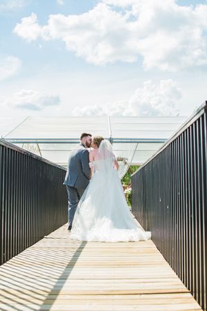 A romantic wedding couple shares intimate moments on a wooden boardwalk with black railings against a cloudy sky.