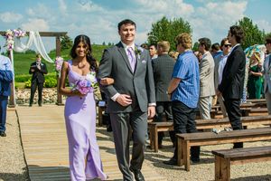 Wedding party members in purple and gray attire walk down an outdoor aisle.