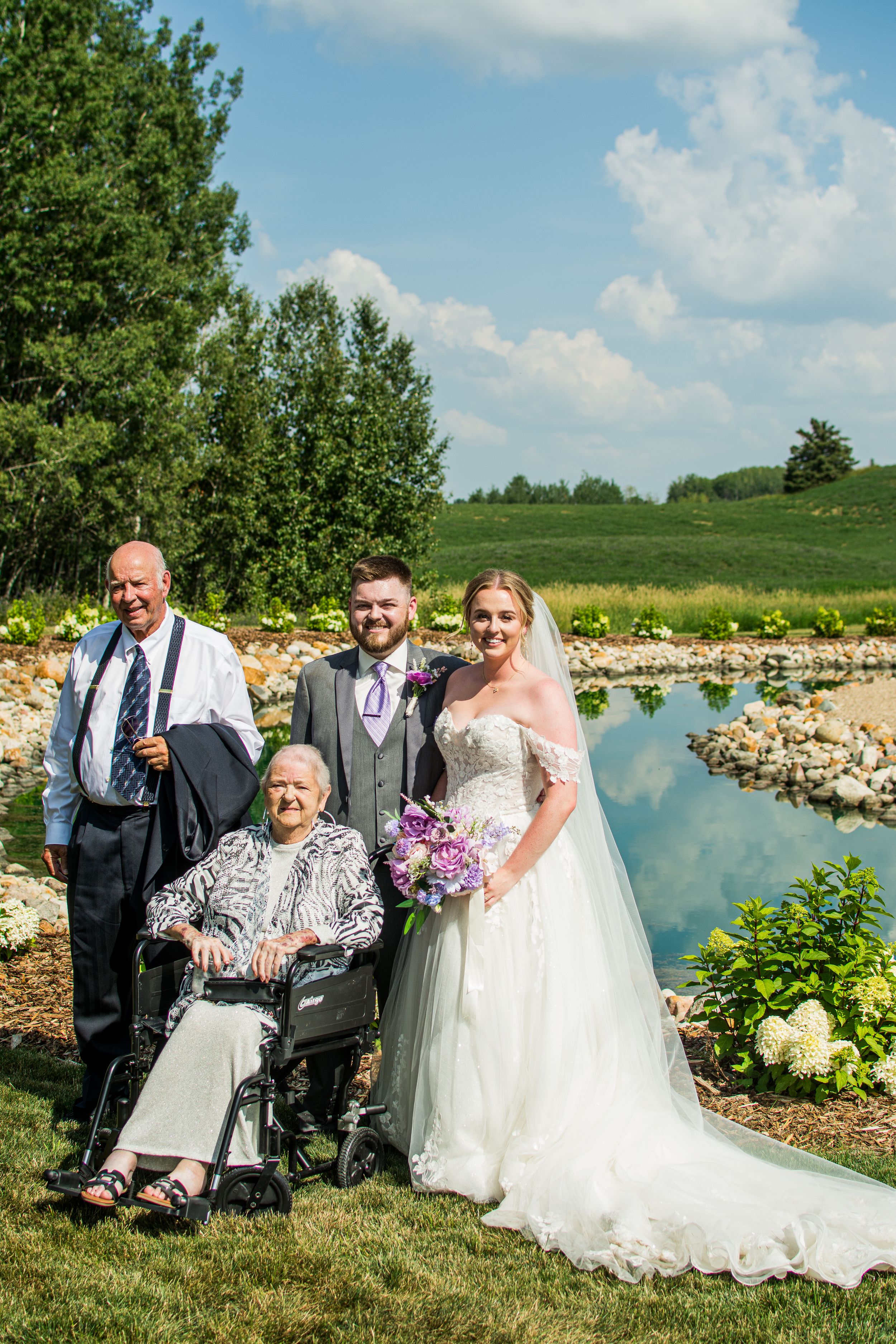 Wedding party members gather for formal photos beside a picturesque pond on a sunny summer day.
