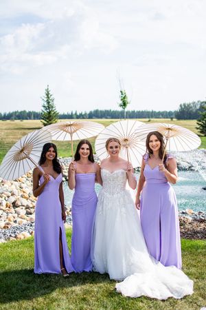 Wedding party holding white parasols posing together by the water in purple and white dresses.