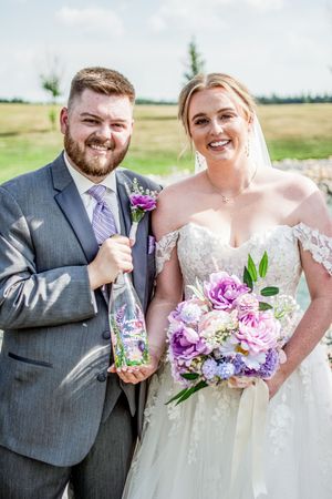 A wedding couple poses outdoors with a purple floral bouquet and decorated bottle on a sunny day.