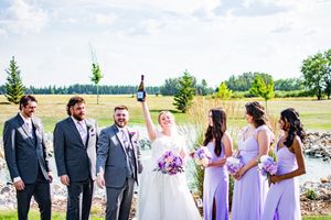 Wedding party members in purple and gray attire pose together outdoors under blue skies.