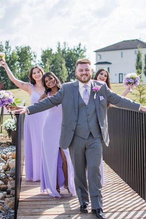 Wedding party in gray suits and purple dresses poses joyfully on a wooden deck.