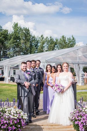 Wedding party posed outdoors with purple floral decorations and white tent backdrop.