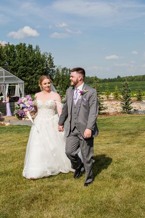 Newlyweds walk hand in hand across a sunny meadow during their summer wedding celebration.