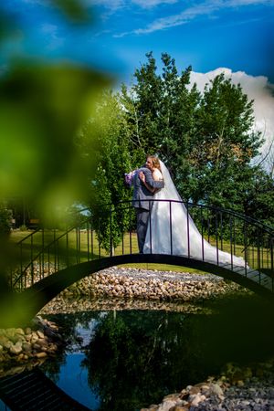 Artistic wedding photo on a bridge over water with trees and rocks in the background.