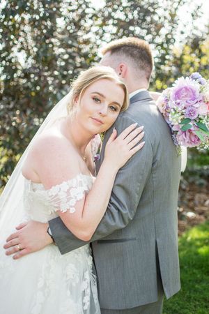 A bride in a lace wedding gown and groom in grey suit embrace during their outdoor wedding photoshoot.