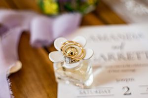 A perfume bottle with a decorative white flower top sits on a wedding invitation on a wooden surface.