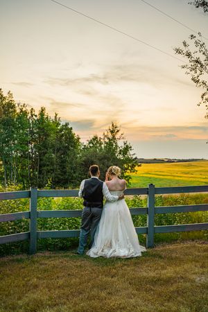 Sunset wedding photos of a couple sitting on a wooden fence overlooking scenic countryside fields.