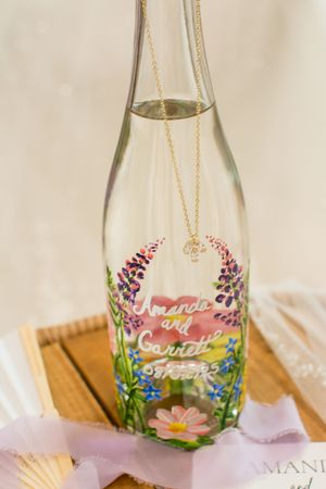 A decorative glass bottle with floral designs and text painted on it sits on a wooden surface.