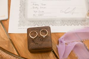 Wedding rings displayed in a wooden box next to marriage documents on a wooden surface.