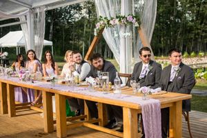 Wedding party seated at a rustic wooden head table decorated with floral arrangements and draped fabric.