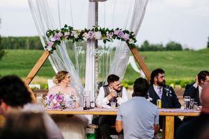 Wedding reception setup with white drapes, floral arrangements and rustic wooden tables on an outdoor deck.