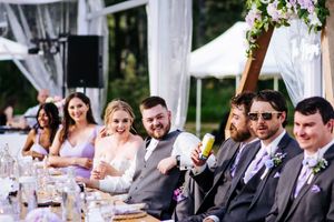 Wedding party seated at long tables under clear tent with purple floral decorations.