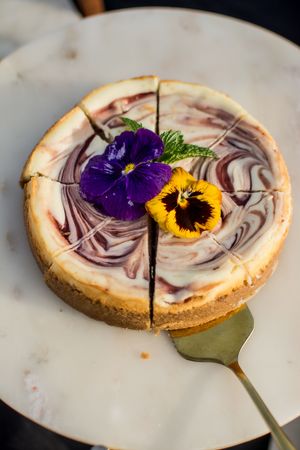 Decorative cheesecake topped with purple pansies and yellow sunflowers on a white marble surface.