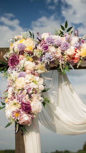 Wedding arch decorated with pink, purple and white flowers with flowing white fabric against a blue sky.
