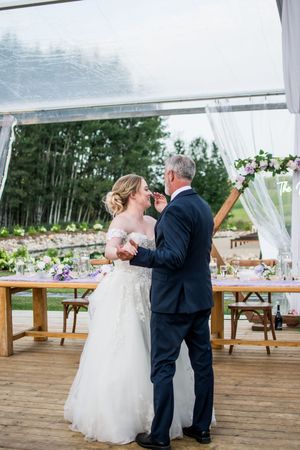 A wedding couple shares their first dance under a white tent on a rustic wooden deck.