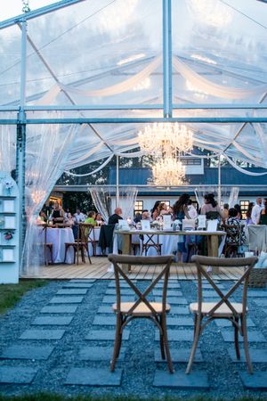 Clear tent wedding venue with elegant chandeliers and rustic wooden cross-back chairs arranged for dining.