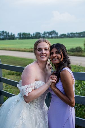 Wedding party members in purple bridesmaid dresses and white gown posing by a fence in a rural setting.