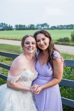 Wedding party in purple and white dresses poses along rustic fence with countryside backdrop.