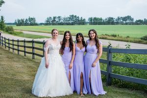 Bridal party in lavender dresses gathers for photos along a rustic fence with rolling fields behind them.