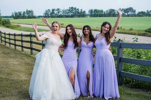Bridesmaids in flowing lavender dresses pose together on scenic farm property.