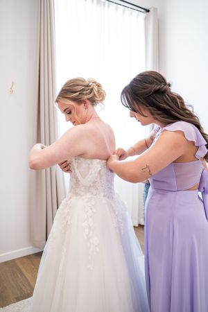 Bridesmaids in lavender dresses help the bride get into her white lace wedding gown before the ceremony.
