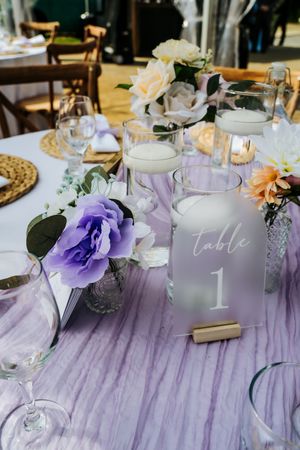 Wedding reception table decorated with purple flowers and frosted glass table number.
