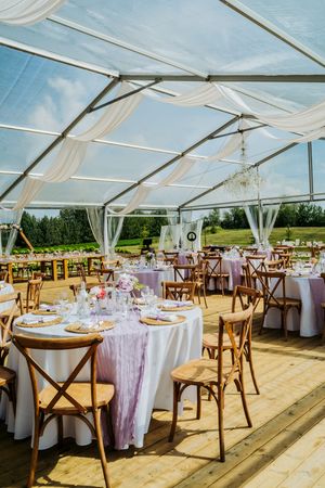 Elegant outdoor wedding reception setup with clear tent, wooden cross-back chairs, and round tables with lavender linens.