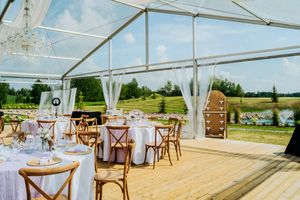 Elegant outdoor wedding reception setup with white tent, crystal chandeliers, and wooden cross-back chairs on a deck.