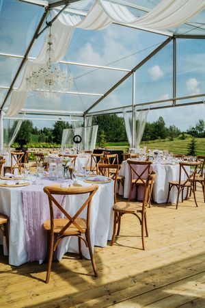 Clear tent reception setup with wooden cross-back chairs and white linens on a wooden deck.