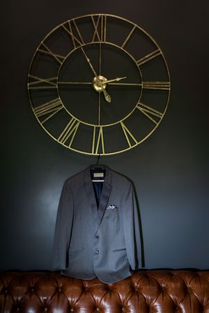 A grey suit jacket hangs beneath a large decorative gold Roman numeral wall clock on a dark wall.