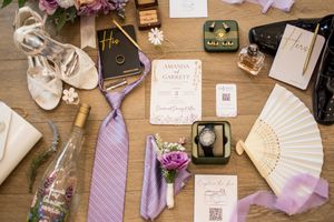 Wedding details flatlay with shoes, tie, fan, and invitation in purple and white tones on wooden surface.