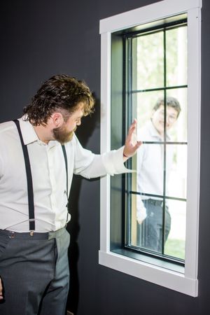 Groomsman in white shirt and suspenders looks out window before wedding ceremony.