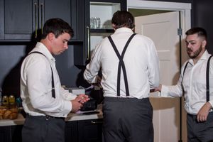 Three groomsmen in white shirts and suspenders getting ready in a dark kitchen before the ceremony.
