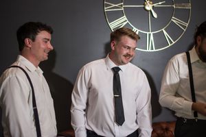 Groomsmen in white shirts and ties getting ready in a dimly lit room with vintage clock on wall.