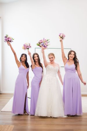 Wedding party in lavender bridesmaid dresses and white bridal gown posing together in a bright studio setting.