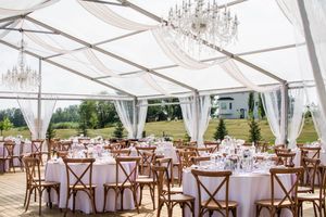 Wedding reception tent setup with elegant chandeliers and cross-back chairs at round tables.