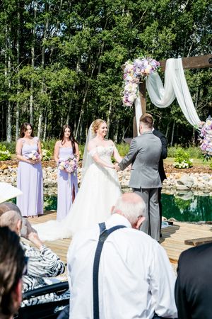 Wedding ceremony guests seated outdoors with purple floral decorations and white chairs on a wooden deck.