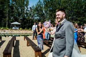 An outdoor wedding celebration with guests seated on wooden benches surrounded by trees.