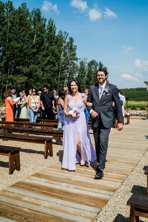 Wedding party walking down wooden aisle at outdoor ceremony under blue sky.
