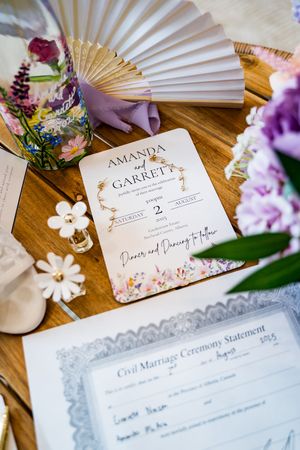 Wedding stationery with floral designs and marriage certificates displayed on a wooden surface with delicate paper fans.