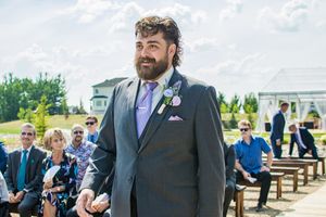 Groom in a dark suit with purple tie awaits at outdoor ceremony venue.
