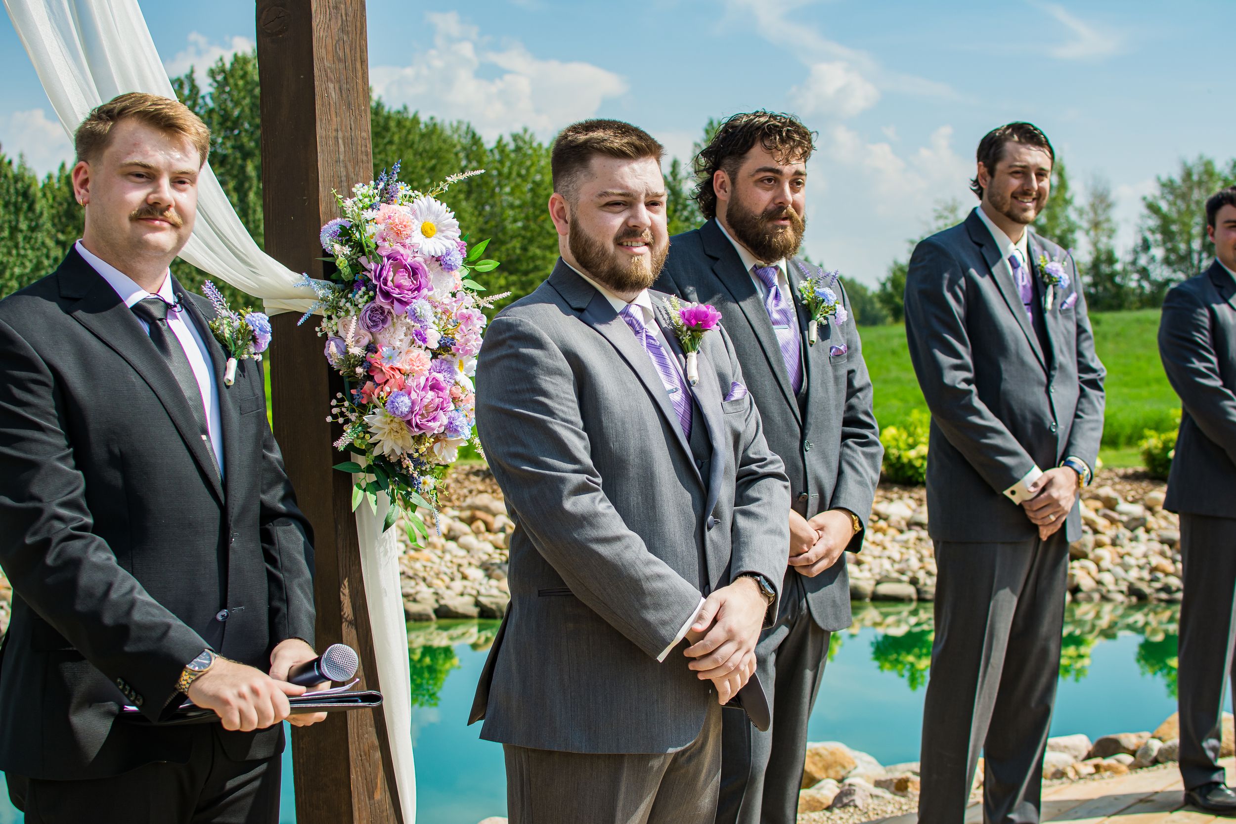 Groomsmen in matching grey suits stand at attention during outdoor lakeside ceremony.