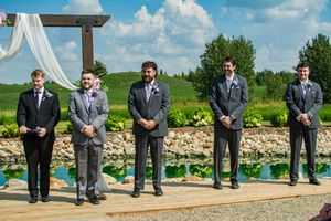 Wedding party members sitting in a row on a dock by a scenic lake with blue skies and greenery in the background.