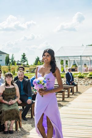 Bridesmaid in flowing lilac dress carries colorful bouquet during outdoor waterfront ceremony.