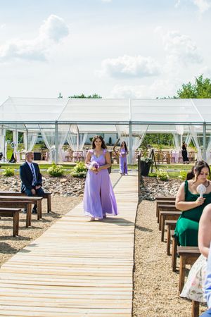 Outdoor wedding ceremony setup with clear tent and purple-dressed attendants walking down wooden aisle.