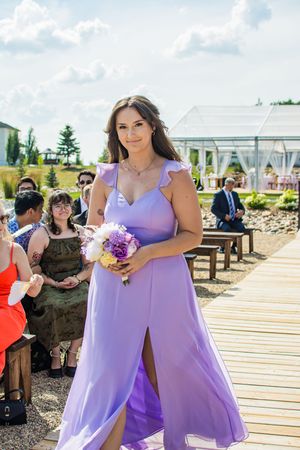 Bridesmaids in flowing lilac dresses walk along a wooden boardwalk at an outdoor wedding venue.