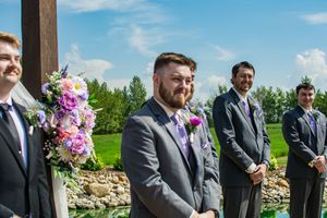 Groomsmen in dark suits stand at outdoor ceremony with floral arrangements and natural backdrop.