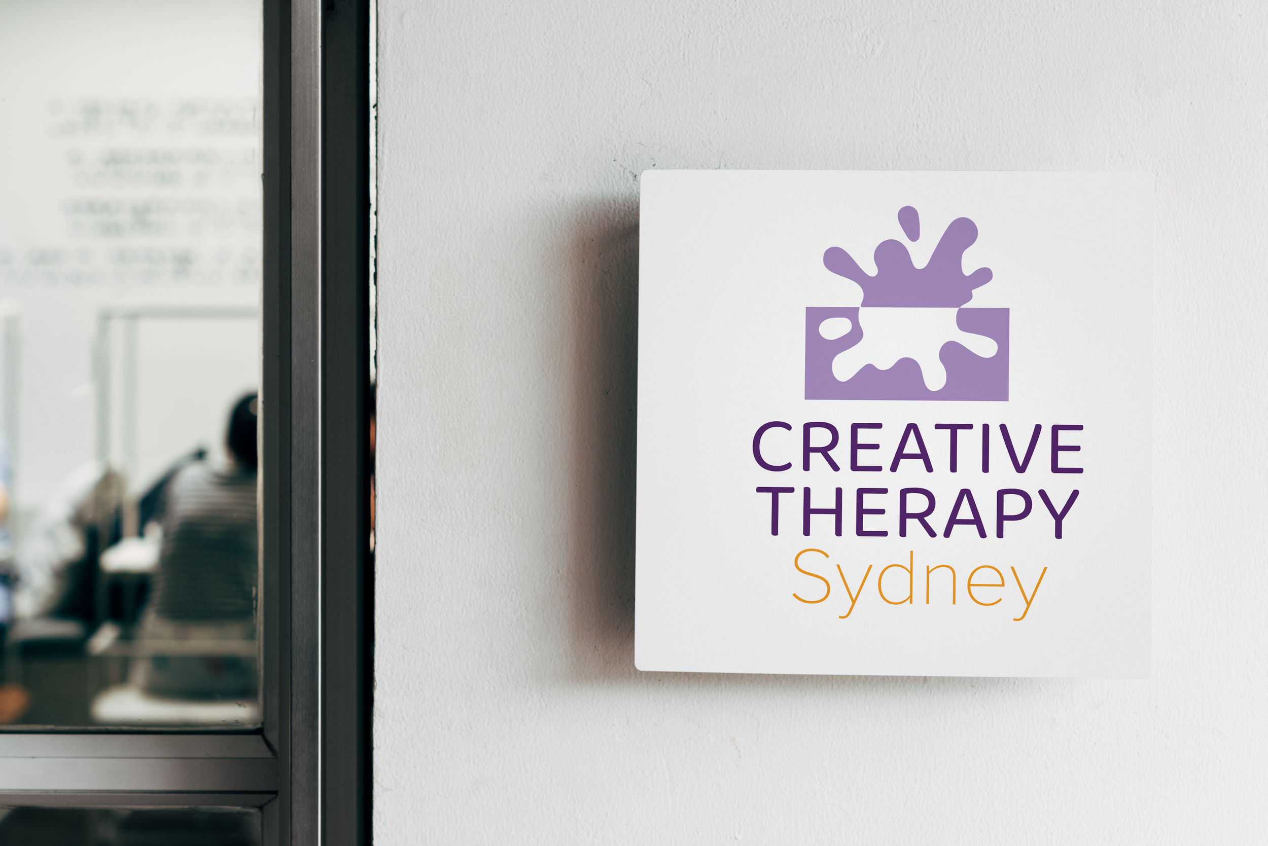 A purple crown logo for Creative Therapy Sydney mounted on a white wall next to a glass window reflection.